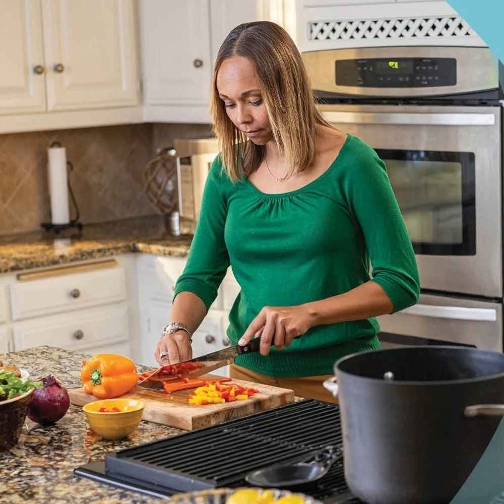 A woman is standing at her kitchen counter cutting up vegetables on a bamboo cutting board